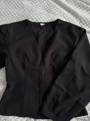 Svart figursydd blus från Nelly - Snygg svart blus från Nelly med figursydd passform och knäppning framtill. Blusen har långa ärmar och djup v-ringning, vilket ger en trendig och stilren look. Perfekt att styla med jeans eller kjol för en chic outfit.