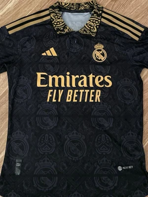Real Madrid tshirt  - Svart Real Madrid tshirt med guldfärgade detaljer. Tryck på framsidan 'Emirates' och Vini Jr #20 på ryggen. Mönstrad krage och axelränder i guld. Mycket bra passform, skön och luftig på sommaren. Storleken är small men e lite mindre i storlek skulle jag säga