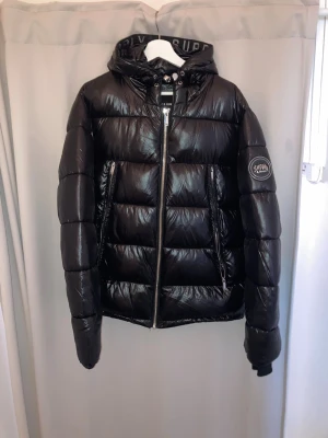 Svart dunjacka från Supply & Demand - Snygg svart dunjacka från Supply & Demand med glansig finish, huva och två dragkedjeförsedda fickor. Jackan har en patch med logga på ärmen och breda kanaler för extra värme. Perfekt för kalla vinterdagar.