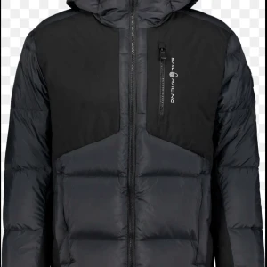 Svart dunjacka från Sail Racing - Snygg svart dunjacka från Sail Racing med huva och dragkedja framtill. Jackan har en bröstficka med dragkedja och logga på vänster sida. Puffig design med paneler och hög krage för extra värme. Perfekt för kalla vinterdagar.