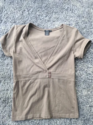 Nougat /  brun/ beige  omlottopp med knappar från Filippa K - Snygg omlottopp från Filippa K med v-ringning och tre dekorativa knappar framtill. Toppen har korta ärmar och är gjord i mjuk bomull, perfekt för en avslappnad och stilren look.
