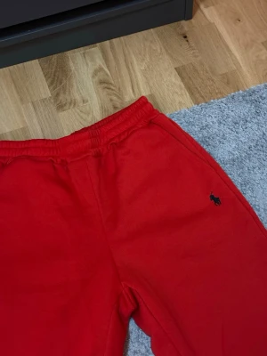 Röda mjukisbyxor från Polo Ralph Lauren - Riktigt snygga röda mjukisbyxor från Polo Ralph Lauren i storlek S, med klassisk logga broderad i mörkblått på ena benet. Byxorna har resår i midjan och sidofickor. Perfekta för chill dagar och streetwear-stil.