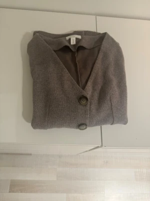 Brun kofta med knappar från H&M - Mysig brun kofta från H&M med fyra stora knappar framtill och två praktiska fickor. Koftan har v-ringning och långa ärmar, perfekt för lager på lager. Materialet är mjukt men ändå stiligt.