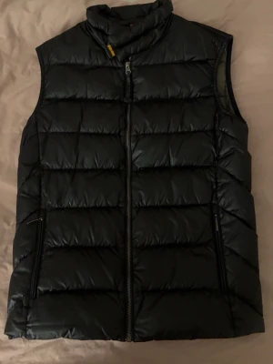 Svart dunväst från Parajumpers - Snygg svart dunväst från Parajumpers, modell Sheen. Västen har hög krage, två dragkedjeförsedda fickor och quiltad design. Insidan är olivgrön med märkets logga och detaljer. Bra passform. Använd ett fåtal gånger.