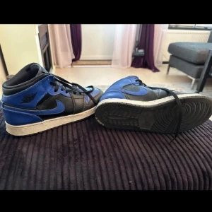 Nike Air Jordan 1 Mid - Snygga Air Jordan 1 Mid sneakers i svart och blått läder med klassisk vit sula och svarta skosnören. Ikonisk Nike Swoosh på sidan och Air Jordan-logga vid ankeln. Perfekta för dig som gillar streetwear och vill sticka ut med en tidlös modell. Pris kan sänkas