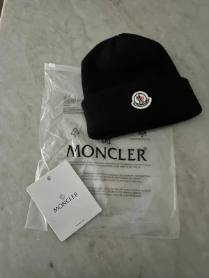 Svart Moncler mössa med logga - Snygg svart mössa från Moncler med uppvikt kant och klassisk Moncler-logga framtill. Tillverkad i mjukt stickat material som håller dig varm och stilren under kalla dagar. Perfekt accessoar för dig som vill ha en clean och exklusiv look.