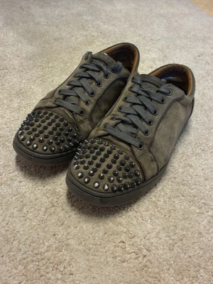 Bruna sneakers med nitar från Louboutin - Unika bruna sneakers från Christian Louboutin med grå snörning och coola svarta nitar över tån. Skorna har klassisk röd sula och är tillverkade i mocka för en lyxig känsla. Perfekta för dig som vill sticka ut med din stil.