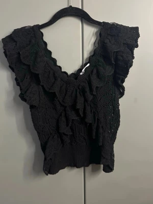 Svart virkad volangblus - Supersöt svart virkad blus med volanger och v-ringning. Toppen är ärmlös och har breda axelband med extra volangdetaljer. Perfekt för dig som vill ha en feminin och trendig look. Materialet känns luftigt och toppen är croppad i modellen.