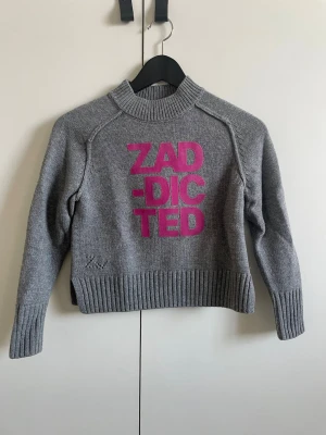 Grå stickad tröja Zadig & Voltaire - Grå stickad tröja från Zadig & Voltaire med hög krage och ribbade muddar. Framtill står det 'ZAD-DICTED' i stor rosa text. Tröjan har en avslappnad passform och är perfekt för dig som gillar statement-plagg. Och även knappt använd