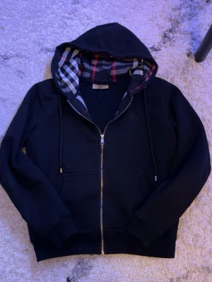 Svart hoodie från Burberry med dragkedja - Svart hoodie från Burberry med hel dragkedja och klassiskt rutigt foder i huvan. Tröjan har snörning i huvan, ribbade muddar och fickor framtill. Perfekt för en stilren och bekväm look. 