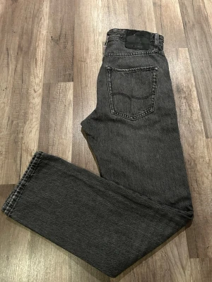 Svart raka jeans från Jack & Jones  - Säljer ett par svarta jeans från Jack & Jones med rak passform och klassisk femficksdesign. Jeansen har en snygg tvättad look och är tillverkade i bomull. Perfekta för en stilren och avslappnad outfit. Storlek W28 L32