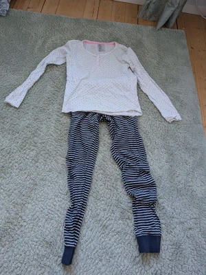 Prickig pyjamas med randiga byxor - Mysig pyjamas med vit långärmad topp med små svarta prickar och rosa kant vid halsen. Byxorna är blå med vita ränder och har muddar vid bensluten. Perfekt för slappa kvällar hemma.