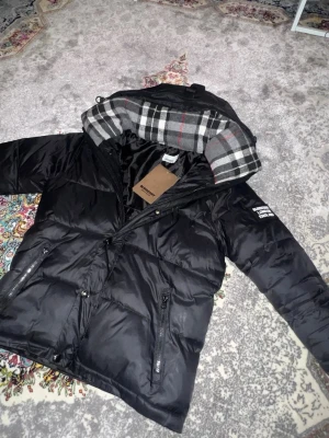 Svart dunjacka från burberry  - Säljer en svart pufferjacka från Burberry med klassiskt rutigt foder i huvan.  Vill bli av med den så fort som möjligt, pris kan diskuteras!