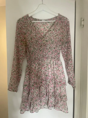 Blommig kortklänning från Zara - Supersöt kortklänning från Zara med rosa och grönt blommönster på vit botten. Klänningen har lång ärm, smock i midjan och volangdetaljer i kjolen. Perfekt för vår och sommar, och har en feminin v-ringning med knappar framtill.