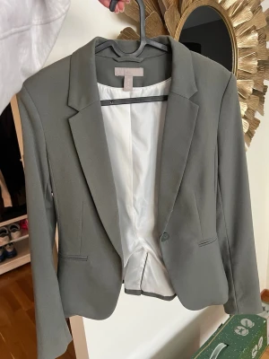 Grön kavaj med klassisk passform - Stilren grön kavaj med klassisk krage och en knapp framtill. Kavajen har två fickor och är helfodrad med vitt tyg på insidan. Snygg, enkel och passar perfekt till många olika looks.