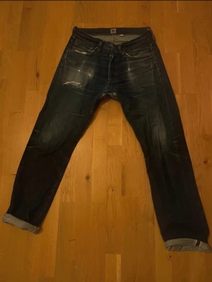 Indigofera blå slitna jeans - Säljer ett par blå jeans från Indigofera med slitningar och hål på bakfickan. Jeansen har raka ben och klassisk femficksdesign. Materialet är kraftig denim i bomull och de har en cool vintagekänsla med tydliga slitdetaljer.