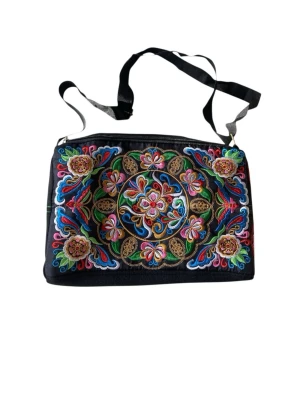 Svart axelväska med färgglad brodyr - Unik svart axelväska med färgglad, detaljerad brodyr i blommönster. Väskan har justerbar axelrem och dragkedja upptill. Materialet är tyg och broderiet går i rosa, blått, grönt, gult och rött. Perfekt för dig som gillar bohemisk och färgstark stil.