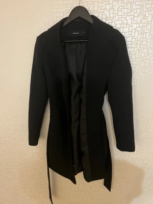 Svart kappa från Vero Moda - Stilren svart kappa från Vero Moda med klassisk krage och knytband i midjan. Kappan har slät yta och är helfodrad med svart tyg på insidan. Perfekt till Hösten, kappan är mer till knäna för mig som är 165cm lång. Inga defekter den är bara använd fåtal gånger 