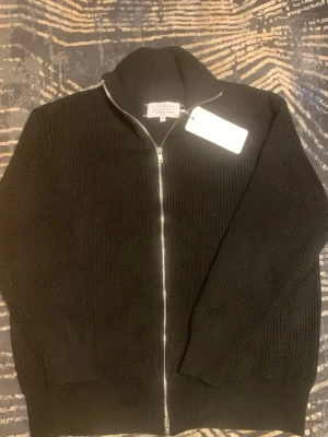 Svart stickad ziptröja Maison Margiela - Säljer en svart stickad ziptröja från Maison Margiela. Tröjan har hög krage, grovstickad struktur och dragkedja framtill. Perfekt för lager på lager och har en clean, minimalistisk look. Märkeslappen syns tydligt i nacken.