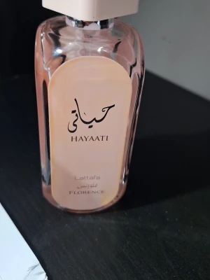 Lattafa Hayaati Florence parfym - Lattafa Hayaati Florence är en parfym i en elegant, rektangulär glasflaska med ljusrosa toner och vit fyrkantig kork. Flaskan har stilren design med arabiska och engelska texter på framsidan. Perfekt för dig som gillar exklusiva dofter. Läcker lite , några droppar liggandes därav priset 