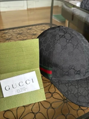 Svart keps från Gucci med GG-mönster - Snygg svart keps från Gucci med klassiskt GG-monogram över hela kepsen. Kepsen har en böjd skärm och detaljer med grönt och rött band på sidan. Tillverkad i canvasmaterial som ger en exklusiv känsla. Perfekt för dig som vill sticka ut med en lyxig accessoar.