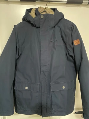 Mörkblå parkas från Tenson - Mörkblå parkas från Tenson med mysigt beige teddyfoder och huva. Jackan har två stora fickor framtill med tryckknappar och en patch på ärmen. Perfekt för kalla dagar och outdoor-äventyr.