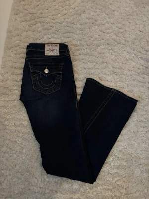 Mörkblå True Religion jeans bootcut - Säljer min tjejs as snygga mörkblå jeans från True Religion med bootcut-modell och ikonisk kontrastsöm på bakfickan. Klassisk femficksdesign och silverfärgad knappdetalj. Perfekta för dig som gillar streetstyle och vill sticka ut med ett par trendiga jeans.