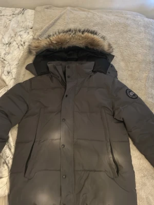 Grå dunjacka från Canada Goose - Säljer en grå dunjacka från Canada Goose med svart logotyp på ärmen och stor huva med fluffig fuskpäls. Jackan har raka fickor och stängs med både dragkedja och knappar. Perfekt för kalla vinterdagar.