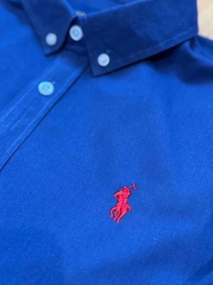 Blå skjorta från Ralph Lauren - Klassisk blå skjorta från Ralph Lauren med röd broderad logga på bröstet. Skjortan har knappar framtill och på kragen samt långa ärmar. Tillverkad i mjuk bomull och passar perfekt till en avslappnad men ändå stilren look.