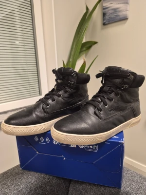 Svarta Björn Borg T1400 HGH TMP high-top sneakers.  - Snygga svarta höga sneakers från Björn Borg i läder med vita gummisulor. Skorna har snörning hela vägen upp och metallöglor upptill för extra streetkänsla. Perfekta för dig som gillar en stilren och urban look.