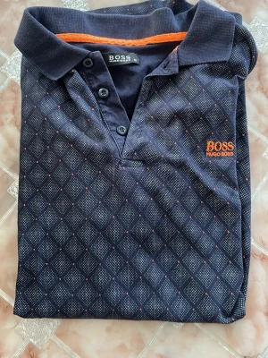 Mönstrad pikétröja från Hugo Boss - Snygg mörkblå pikétröja från Hugo Boss med diskret rutmönster och orange detaljer. Tröjan har klassisk krage, knappar framtill och broderad logga på bröstet. Perfekt för en stilren och avslappnad look.
