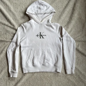 Vit hoodie från Calvin Klein Jeans - Säljer en stilren vit hoodie från Calvin Klein Jeans med deras ikoniska logga i silver på bröstet. Tröjan har en klassisk känguruficka och ribbade muddar vid ärmslut. Perfekt för en avslappnad look.