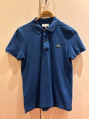 Blå pikétröja från Lacoste - Klassisk blå pikétröja från Lacoste med korta ärmar och krage. Tröjan har två knappar framtill och den ikoniska krokodilloggan på bröstet. Tillverkad i bomull för en bekväm och luftig känsla.