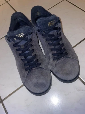 Grå mockasneakers från Polo Ralph Lauren - Snygga grå sneakers från Polo Ralph Lauren i mjuk mocka med svarta snören och diskret logga på plösen. Skorna har en klassisk siluett och svart sula som ger en clean look. Perfekta för dig som gillar stilrena och tidlösa sneakers.