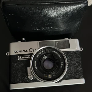 Konica C35 analog kamera med väska - Säljer en klassisk Konica C35 analog kamera i silver och svart med Hexanon 38mm f/2.8-objektiv. Kameran har retro design med metallhölje och detaljer i svart läderimitation. Praktisk och smidig storlek, perfekt för dig som gillar vintagefoto. Medföljer original svart kameraväska.