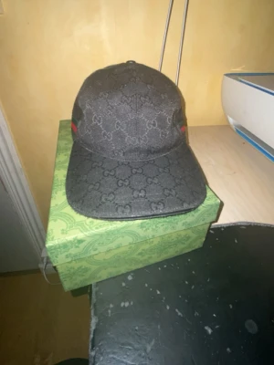 Svart Gucci keps med GG-mönster - Box kommer med! Snygg svart keps från Gucci med klassiskt GG-monogram över hela kepsen. På sidan finns ett grönt och rött band som ger den en ikonisk touch. Kepsen har böjd skärm och justerbar passform. Materialet ser ut att vara canvas med detaljer i tyg.