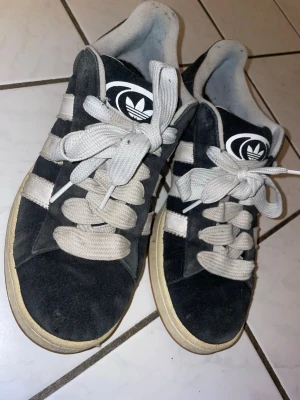 Adidas campus-svarta - Klassiska svarta Adidas sneakers med de ikoniska tre vita ränderna på sidorna och vit Adidas-logga på plösen. Skorna har vita snören och en beige sula. Ovandelen är i mockaliknande material och insidan är ljusgrå. Perfekta för en sportig och avslappnad stil.