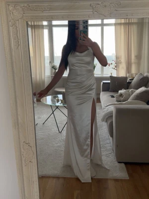 Vit långklänning med slits - Elegant vit långklänning i satinliknande material med smala axelband och en hög slits på ena sidan. Klänningen har en figurnära passform och v-ringad halsringning som ger en stilren och modern look.