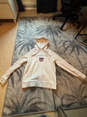 Beige hoodie från GANT med logga - Beige hoodie från GANT med stor broderad logga och sköld på bröstet. Klassisk passform med huva och ribbade muddar vid ärmslut och nederkant. Tillverkad i mjukt bomullsmaterial som känns skönt mot huden. Perfekt för en clean och avslappnad stil.
