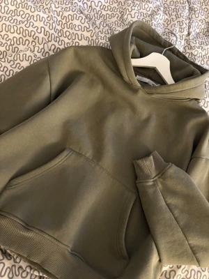 Hoodie - Snygg olivgrön hoodie med klassisk huva och stor magficka framtill. Hoodien har ribbade muddar vid ärmslut och nederkant, samt en relaxed fit som passar perfekt till streetwear-stilen. Tillverkad i mjukt och bekvämt material, troligtvis bomullsblandning.