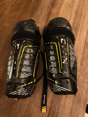CCM Tacks benskydd svart/gul 28 cm - Säljer ett par svarta och gula benskydd från CCM Tacks-serien. Skydden har justerbara remmar och vadderad insida för extra komfort. Ytan har vita och gula detaljer samt grafiskt mönster. Perfekta för hockey eller annan kontaktsport. Passar längd 134-146