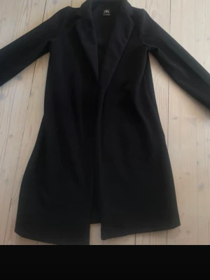 Svart kappa från Zara med krage - Stilren svart kappa från Zara med klassisk krage och rak passform. Kappan är enkel och tidlös, perfekt för dig som gillar minimalistisk stil. Materialet ser ut att vara en mjuk ullblandning som håller dig varm under kyliga dagar.