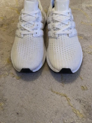 Vita Adidas Ultraboost sneakers - Snygga vita Adidas Ultraboost sneakers med stickad ovandel i mesh för grym komfort och andningsförmåga. Skorna har klassiska tre ränder på sidan, vit sula med Boost-dämpning och svart yttersula. Perfekta för dig som gillar stilrena och sportiga sneakers.