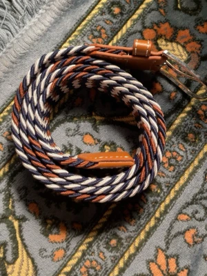 Randigt flätat bälte i blått och brunt - Snyggt flätat bälte med randigt mönster i marinblått, vitt och brunt. Bältet har detaljer i brunt skinn och ett klassiskt metallspänne. Perfekt accessoar för att lyfta din outfit med lite färg och stil.