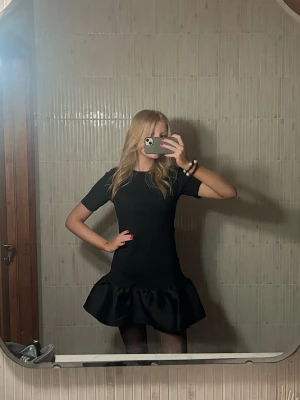 Svart kort klänning med volang - Snygg svart kort klänning med volang nertill och rund hals. Klänningen har korta ärmar och en figurnära passform upptill. Perfekt för dig som gillar en stilren look med en twist. Passar till många olika tillfällen. Endast använd en gång❣️