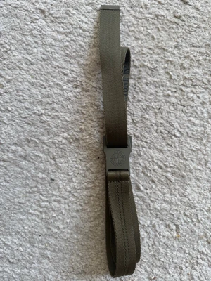 Olivgrönt bälte från Stone Island - Snyggt olivgrönt bälte från Stone Island med klassiskt spänne där loggan är ingraverad. Bältet är gjort i slitstarkt syntetmaterial och har en stilren, minimalistisk design som passar till många olika outfits.