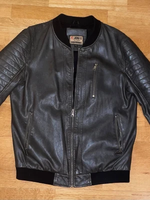 Svart läderjacka - Snygg svart läderjacka från Jerry's med quiltade detaljer på axlarna, tre dragkedjefickor framtill och ribbade muddar vid ärmslut och midja. Jackan har en klassisk bombermodell och svart innerfoder. Perfekt för en cool streetstil.