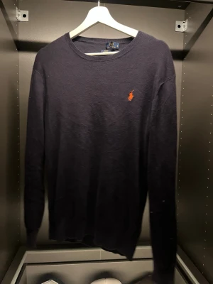 Mörkblå stickad tröja från Polo Ralph Lauren - Mörkblå stickad tröja från Polo Ralph Lauren med klassisk orange broderad logga på bröstet. Tröjan har rund halsringning och långa ärmar. Perfekt för dig som gillar stilrena och tidlösa plagg. Den är använd men har mycke kvar att ge! 8/10