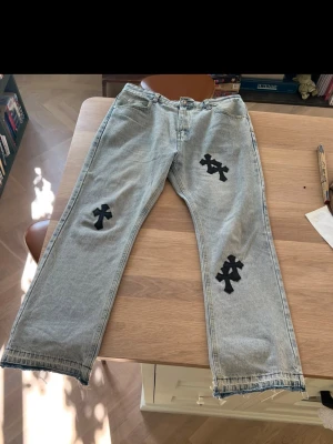 Grå blåa chrome heart jeans med svarta korsdetaljer - Säljer ett par grå  blåa chrome heart jeans med raka ben och unika svarta korsapplikationer på både fram- och baksidan. Jeansen har klassisk femficksdesign och är tillverkade i denim. Perfekt för dig som vill sticka ut med en egen still.