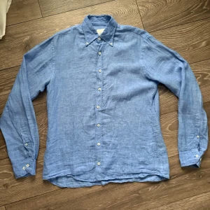 Oscar Jacobson  - Ljusblå skjorta i linne från Oscar Jacobson med klassisk krage och vita knappar hela vägen fram. Skjortan har långa ärmar med knapp vid ärmslut och en avslappnad passform. Perfekt för en chill och stilren look.